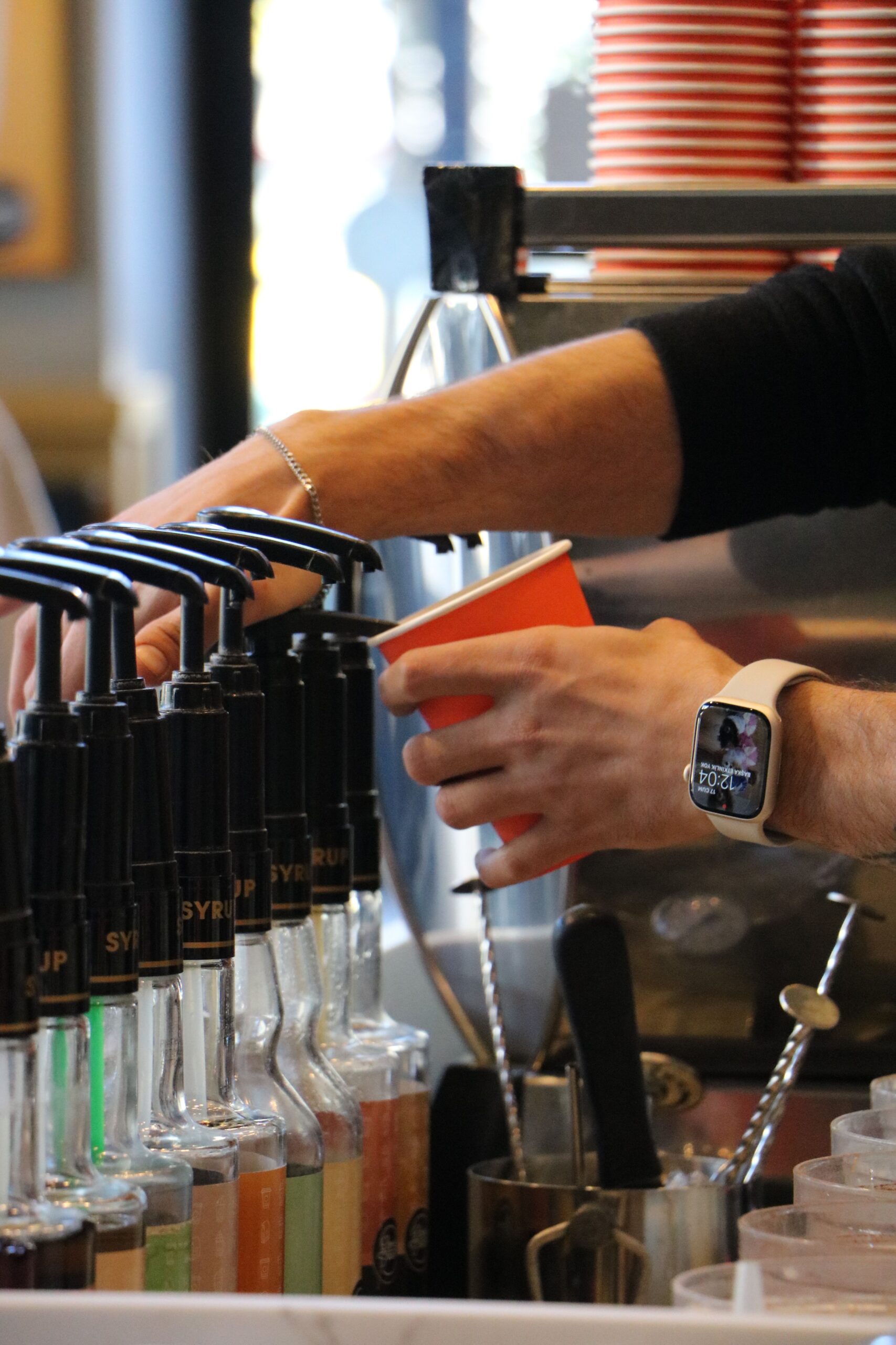 Barista adding flavored syrups to coffee with smartwatch in cafe setting.
